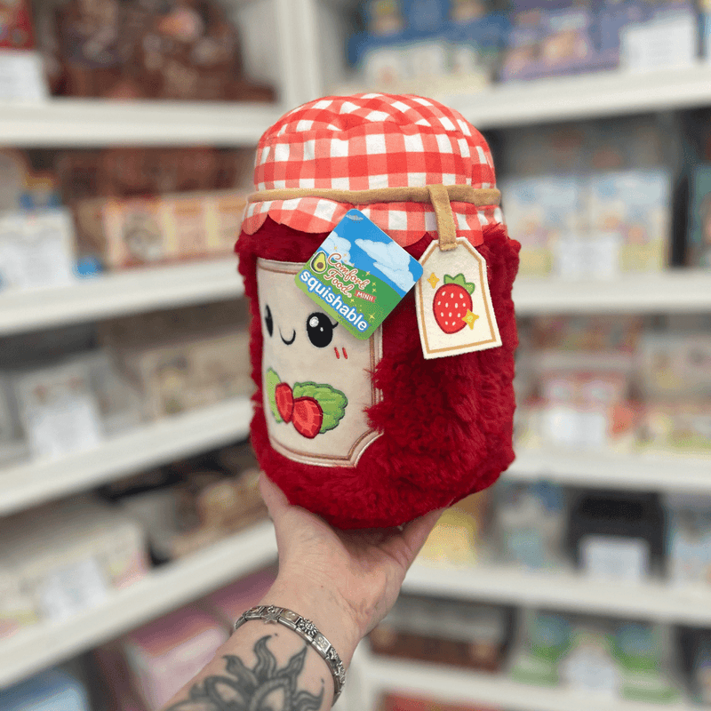 Mini Squishable Comfort Food Strawberry Jam plush toy held in hand, angled view showing the soft red jam jar, gingham lid and strawberry gift tag