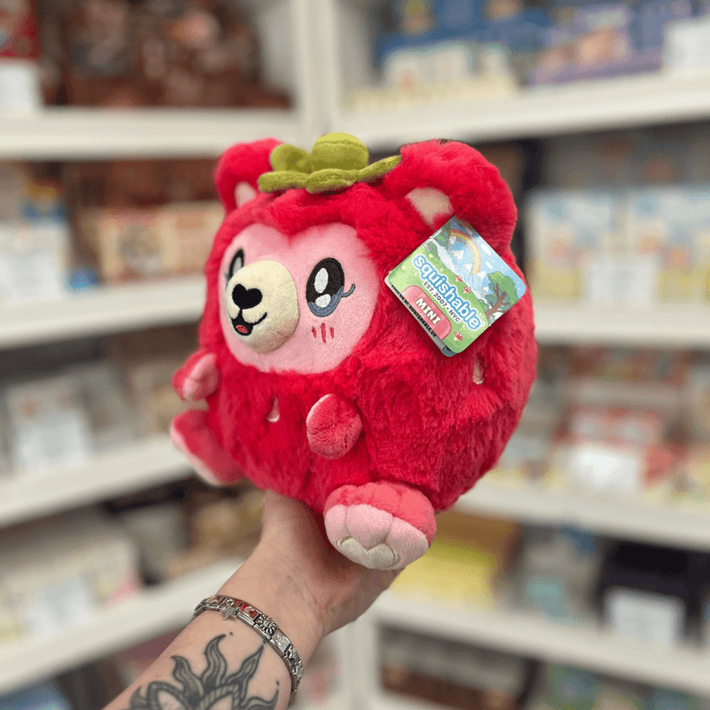 Mini Squishable Strawbeary plush toy held in hand, angled side view showing the soft red plush body, pink paws, embroidered face and green leaf detail on top