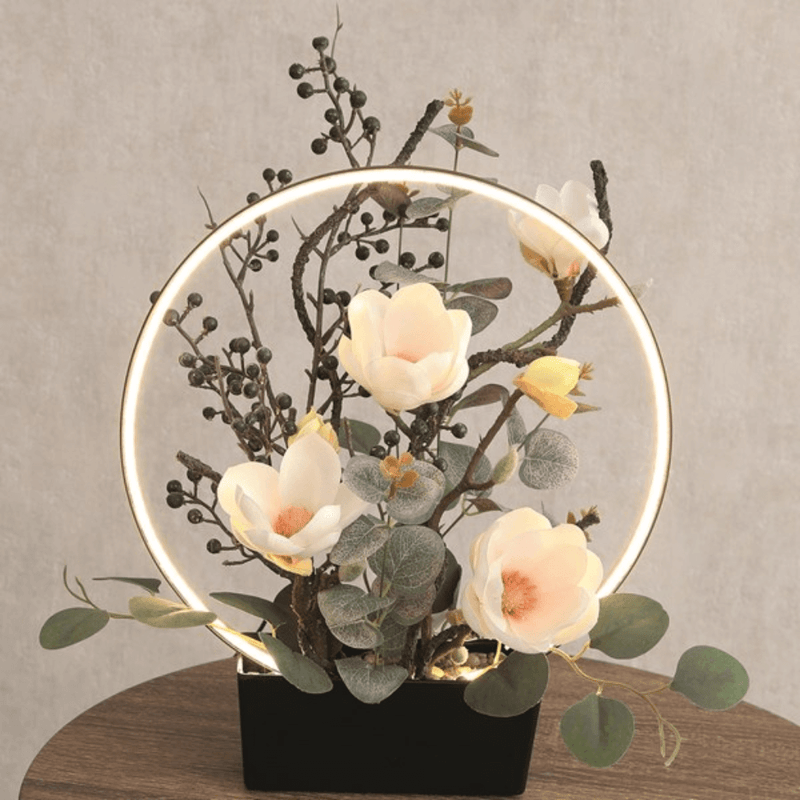 Lifestyle image of the LED floral halo lamp glowing on a side table with white blossoms and foliage.