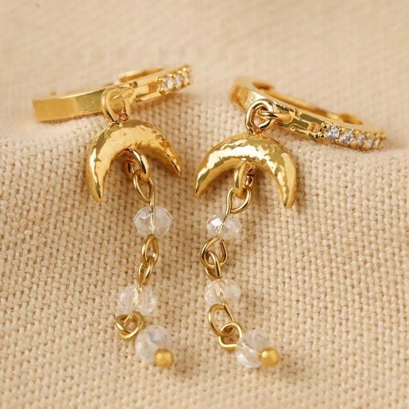 Gold huggie hoop earrings with hammered moon charms and crystal bead chains styled on neutral fabric.