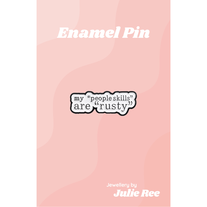 White enamel pin with black text reading my “people skills” are “rusty”, displayed on a pink backing card.