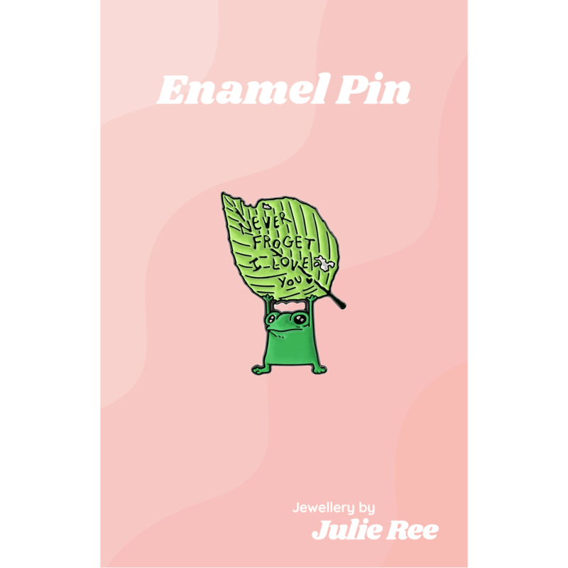 Green enamel pin of a cute frog holding a leaf that says Never Froget I Love You, displayed on a pink backing card.