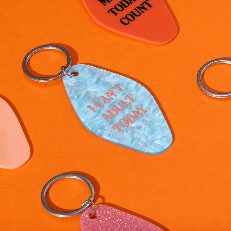 Lifestyle image of Ohh Deer blue pearl-effect keyring with the quote I Can’t Adult Today on orange background.