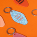 Lifestyle image of Ohh Deer blue pearl-effect keyring with the quote I Can’t Adult Today on orange background.