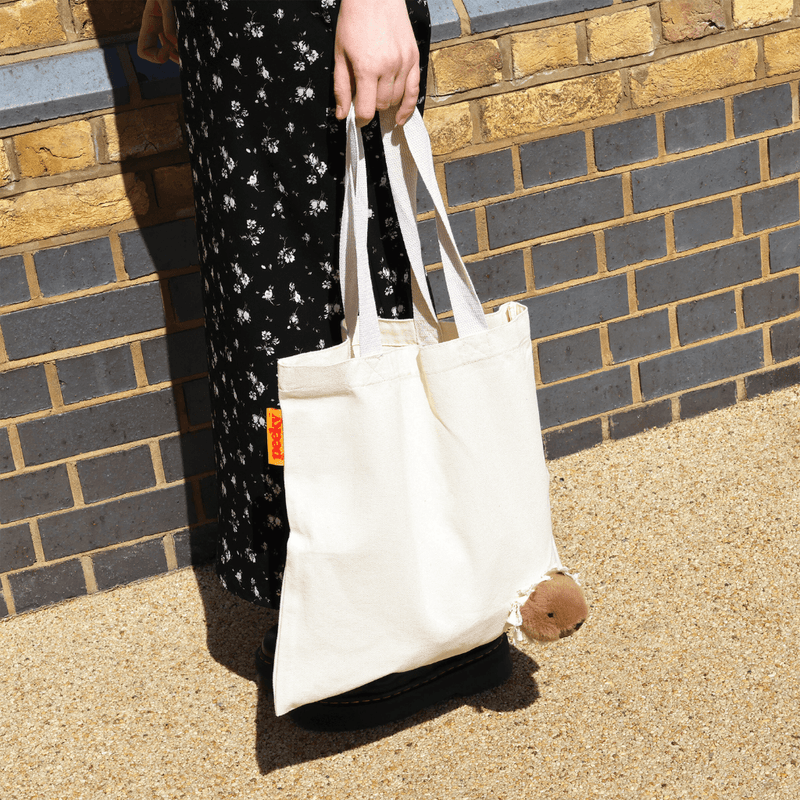 Side view of a cream canvas capybara tote bag with long handles and a plush brown animal head emerging through the fabric.
