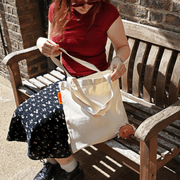 Woman sitting on a wooden bench holding a natural cotton tote with a cute plush capybara peeking through the front.