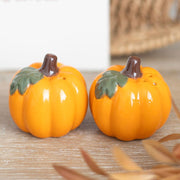 Pair of glossy ceramic pumpkin salt and pepper shakers styled on a wooden table with autumn foliage.
