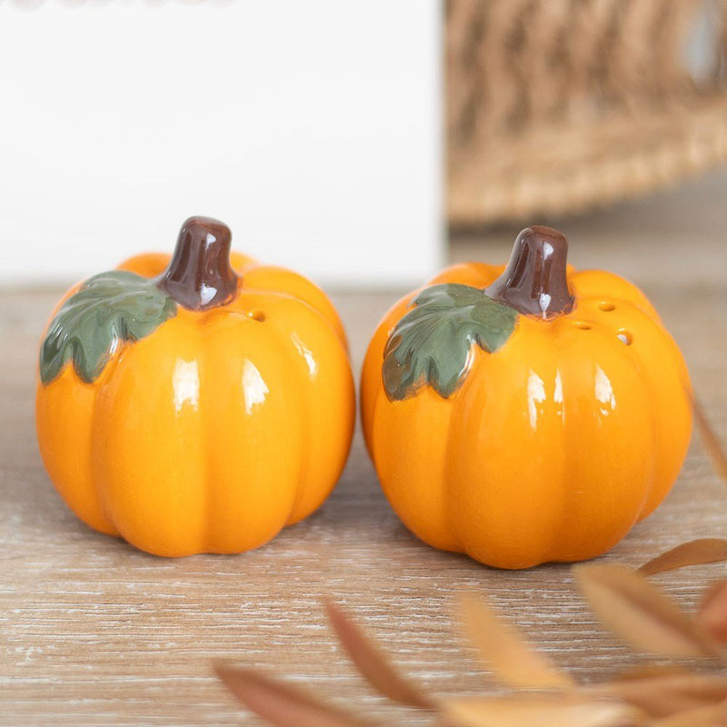 Pair of glossy ceramic pumpkin salt and pepper shakers styled on a wooden table with autumn foliage.

