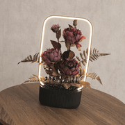 Lifestyle image of the mains-powered LED floral frame lamp glowing on a wooden side table.