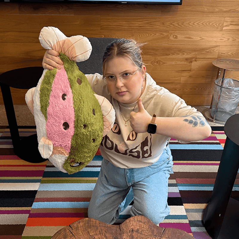 Person holding Squishable Rainbow Trout plush toy in a room with wooden wall and colourful rug.