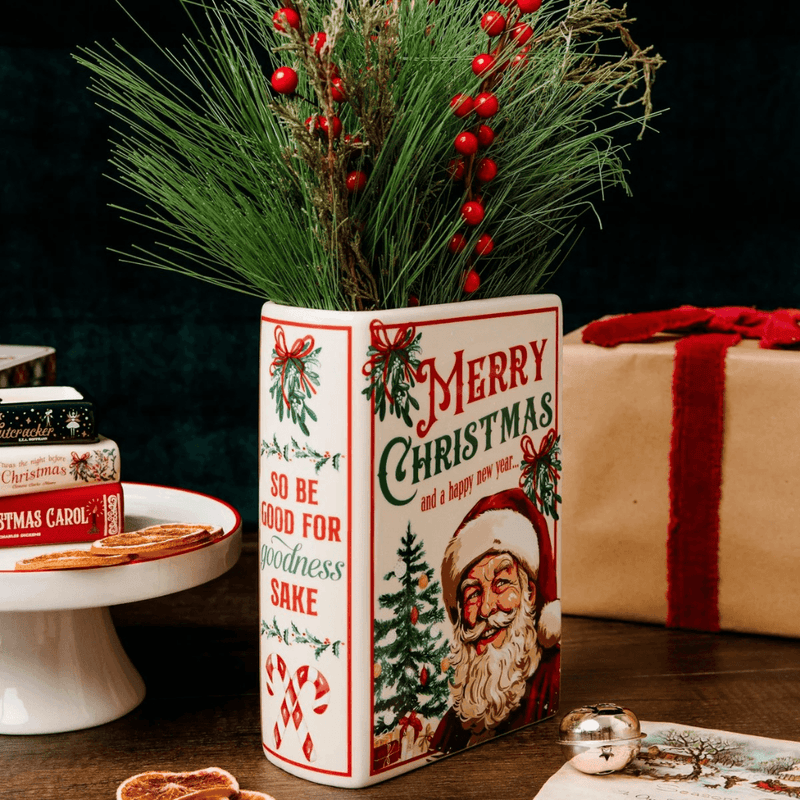 Santa Christmas book vase with winter greenery and red berries styled on a wooden table beside wrapped Christmas presents