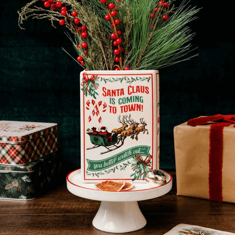 Reverse of Santa ceramic book vase filled with pine branches and red berries, styled with festive gift boxes