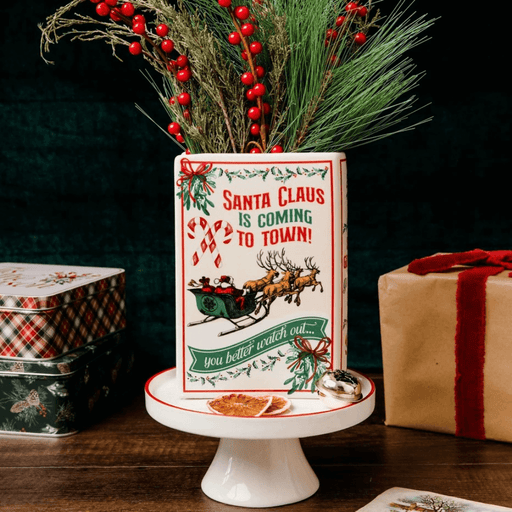 Reverse of Santa ceramic book vase filled with pine branches and red berries, styled with festive gift boxes