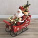 Lifestyle view of musical Santa in sleigh with presents and mini tree glowing on the floor.