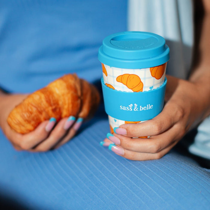 Person holding Sass & Belle croissant travel coffee cup with blue lid and croissant print