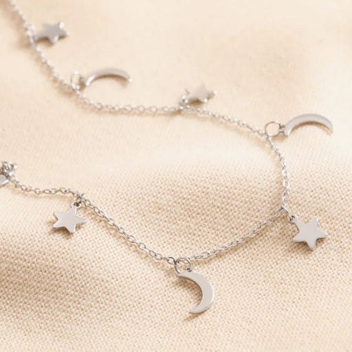 Close-up of a silver stainless steel necklace with small star and crescent moon charms spaced along a fine chain on neutral fabric.