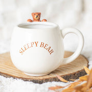 Cream ‘Sleepy Bear’ mug on rustic wood slice with bear figurine peeking over the rim.
