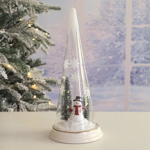 LED snowman glass tree glowing on a table beside a Christmas tree, cosy winter backdrop.