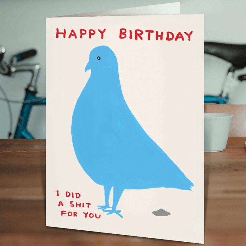 David Shrigley birthday card on a wooden table, blue pigeon artwork and rude caption visible.


