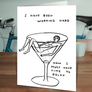 David Shrigley “Working Hard, Time To Relax” card standing on a wooden table in a lifestyle setting.