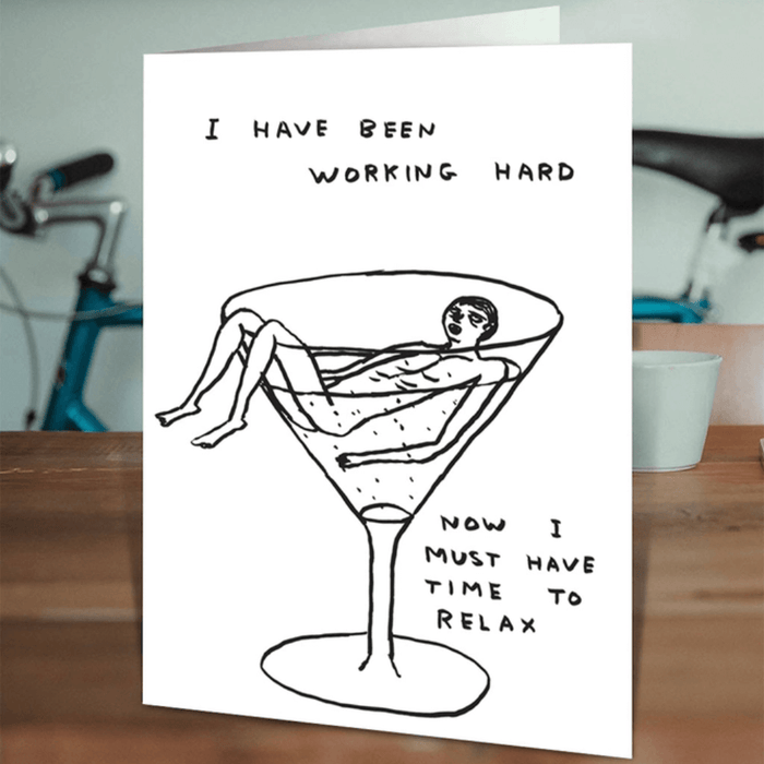David Shrigley “Working Hard, Time To Relax” card standing on a wooden table in a lifestyle setting.