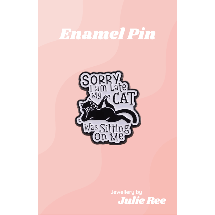 Black and white enamel pin with a lounging cat and text saying Sorry I am late my cat was sitting on me, on a pink backing card.