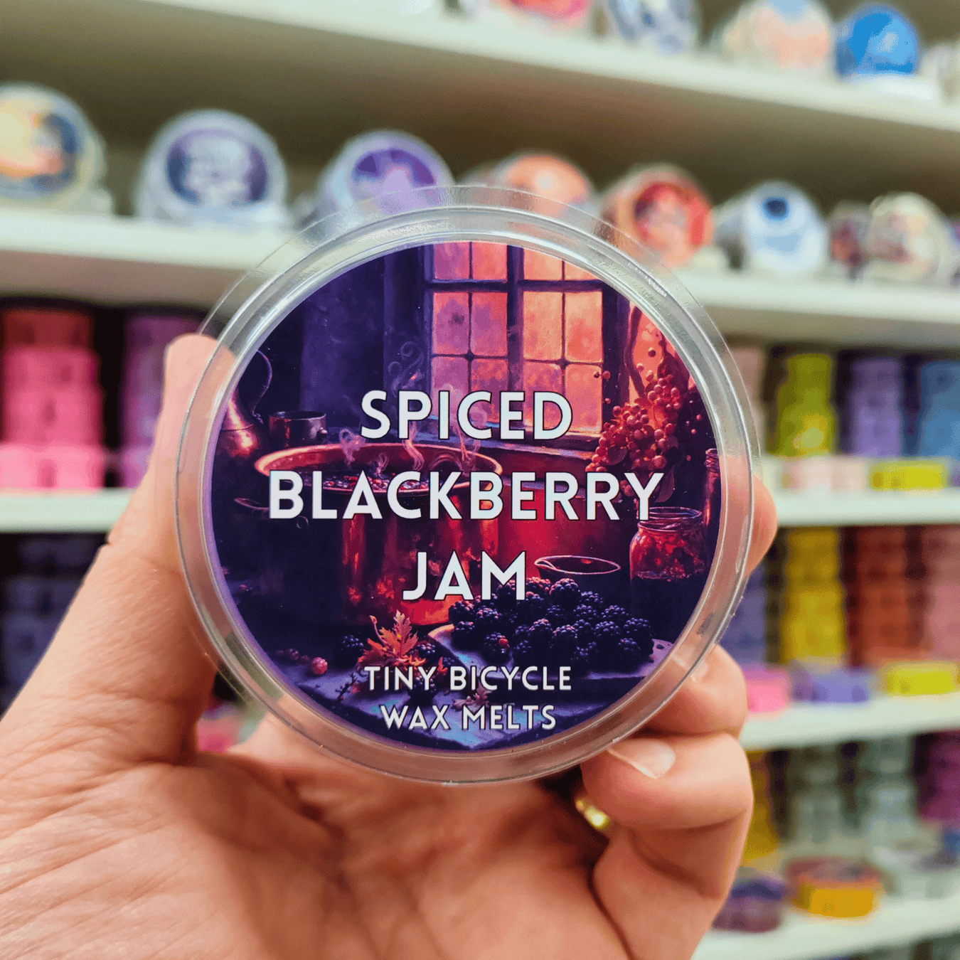 Spiced Blackberry Jam wax melt pot held in hand with illustrated label, lifestyle product image with wax melt shelves behind