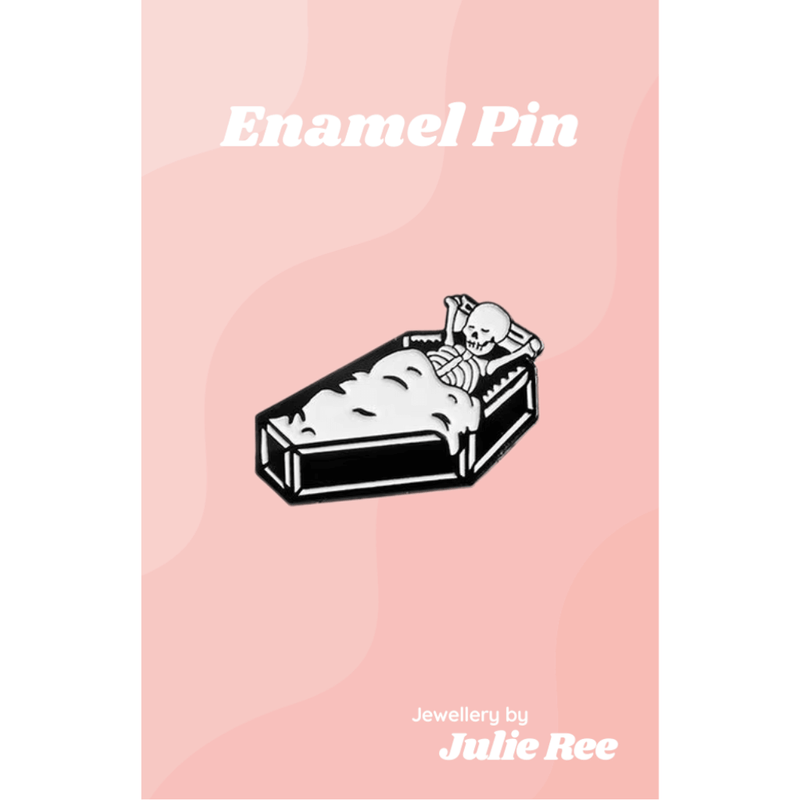 Black and white enamel pin of a relaxed skeleton lying in a coffin with bubble bath, on a pink backing card.