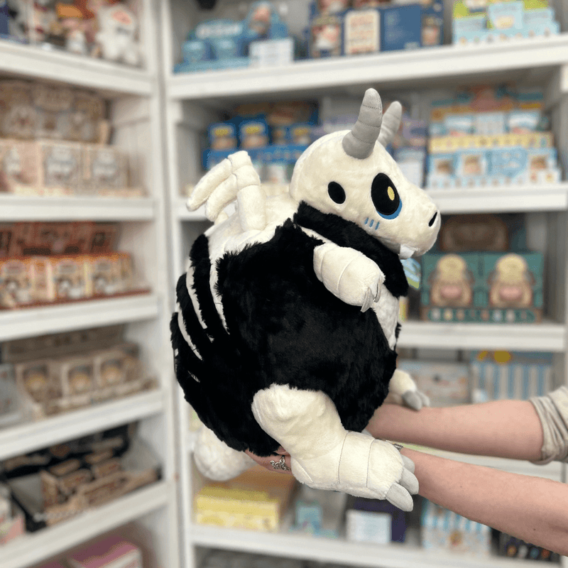 Squishable Bone Dragon plush toy held in hands in a gift shop, side view showing wing and rib detail