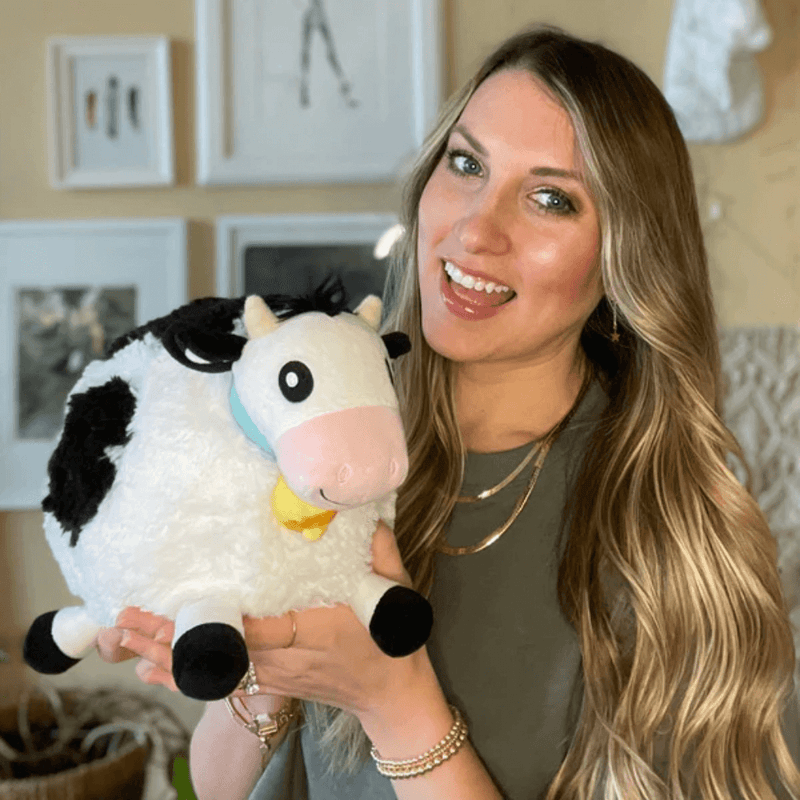 Person holding Squishable Mini Cow plush toy with black and white patches and jingle bell collar