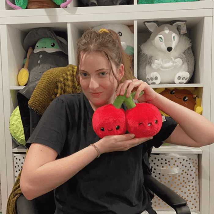 Person holding Squishable Snacker Cherries plush toy in front of a shelf display of plush toys
