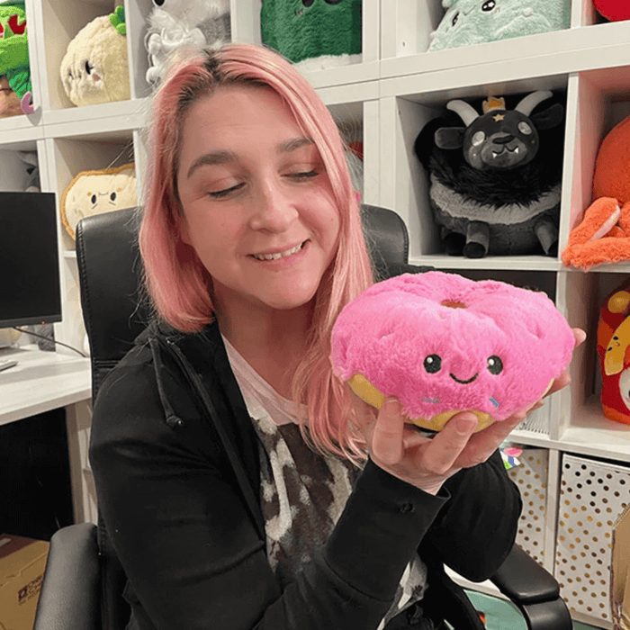Person holding Squishable Snacker Pink Donut plush toy in front of a shelf display of plush toys