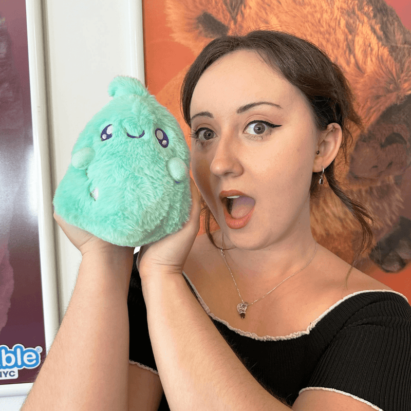 Person holding Squishable Snacker Teal Ghost plush beside their face to show its cuddly size