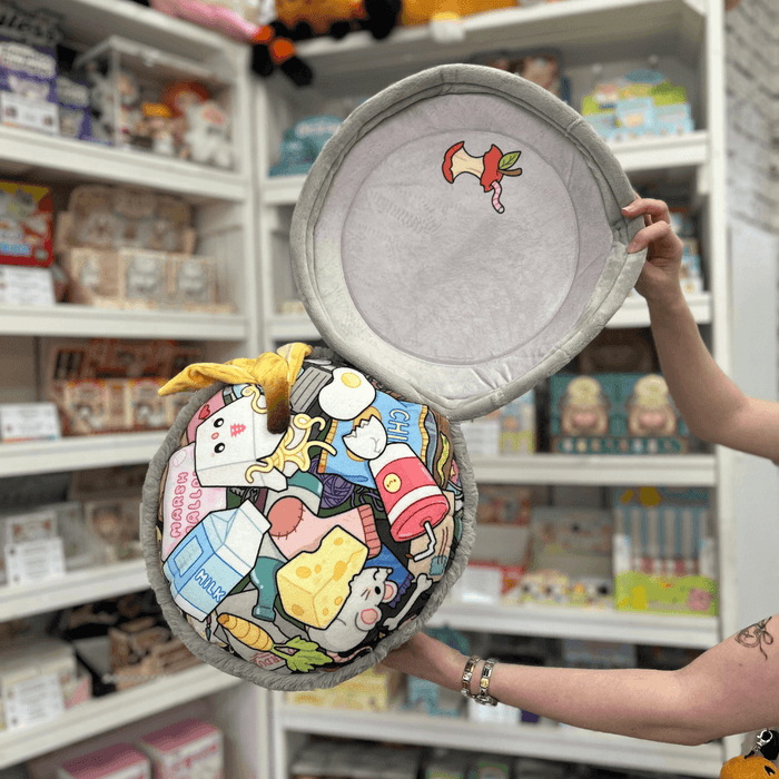 Squishable Trash Can plush toy held in hands in a gift shop, opened to show illustrated rubbish inside