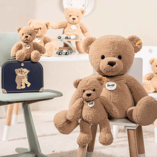 Steiff Ben Teddy Bear in beige teddy fleece displayed in a nursery with other teddy bears and a blue suitcase