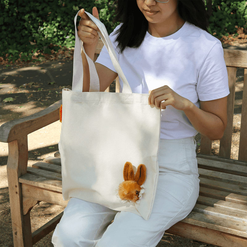 Natural cream canvas shopper with a three-dimensional bunny head, held by a woman sitting on a wooden bench.
