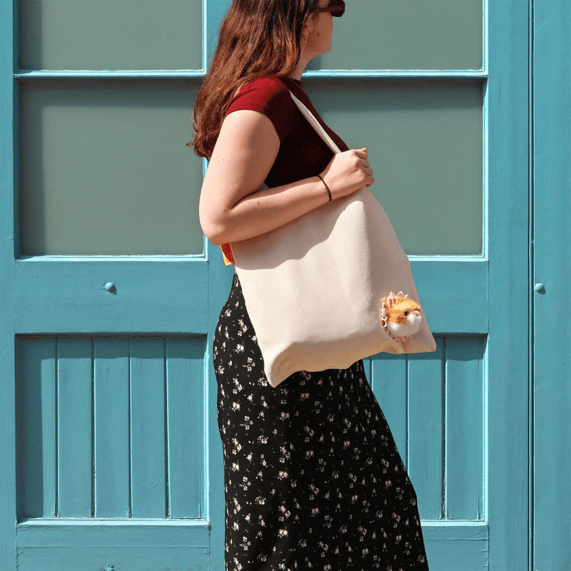 Woman wearing a cream canvas Peeky Cat tote bag over her shoulder beside a turquoise doorway.
