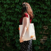 Woman carrying the SUCK UK Peeky Cat natural canvas tote bag with a fluffy ginger-and-white kitten peeking out, against an ivy-covered wall.