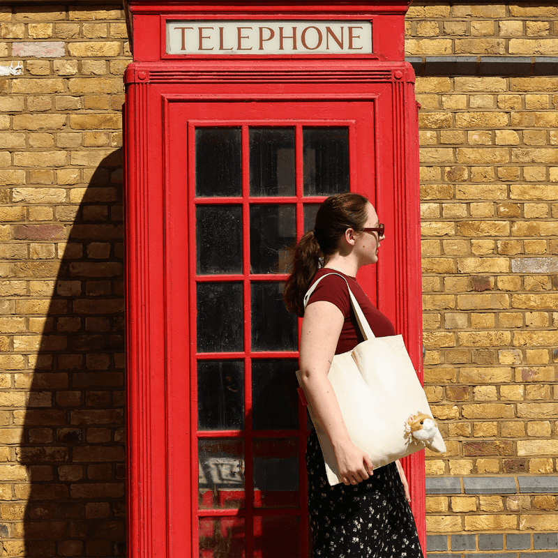 Woman wearing the SUCK UK Peeky Cat tote bag beside a red British telephone box, showing the fluffy cat detail.