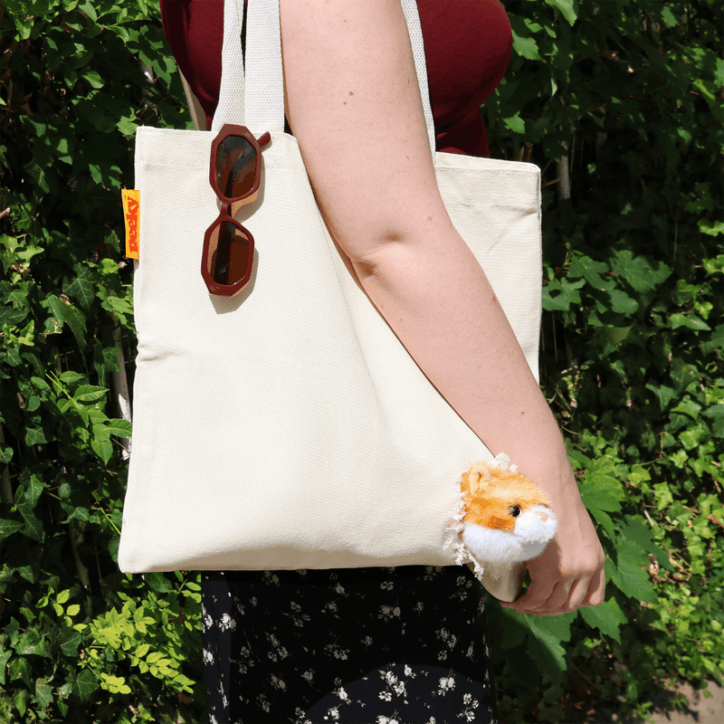 Close-up of a woman carrying the Peeky Cat canvas tote bag over her shoulder, with the kitten head visible against green foliage.