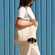Woman wearing the SUCK UK Peeky Dog tote bag over her shoulder against a blue wall, with the plush puppy face visible.