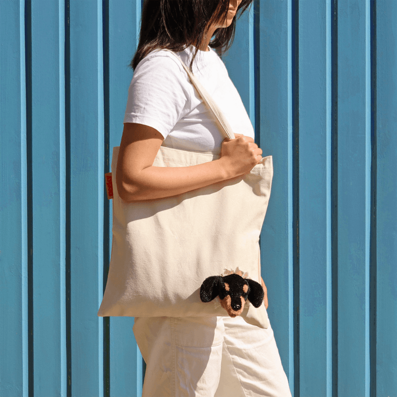 Woman wearing the SUCK UK Peeky Dog tote bag over her shoulder against a blue wall, with the plush puppy face visible.