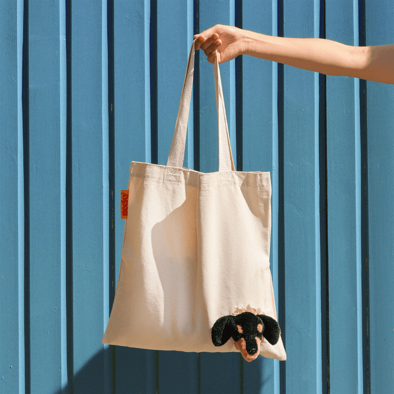Natural cream canvas Peeky Dog tote bag held by its shoulder straps in front of a blue wall, showing the fluffy floppy-eared dog head.