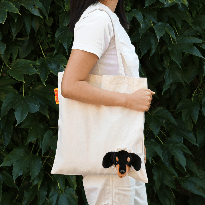 Woman carrying a natural cotton SUCK UK Peeky Dog tote bag with a plush black-and-tan puppy head peeking out, against green ivy.