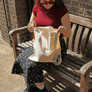 Woman sitting on a wooden bench holding a natural cotton SUCK UK Peeky Donkey tote bag with a fluffy grey donkey head peeking out.