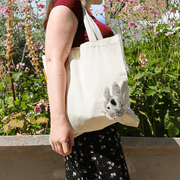 Woman wearing the Peeky Donkey canvas shopper over her shoulder in a flower garden, showing the plush grey donkey's long ears.