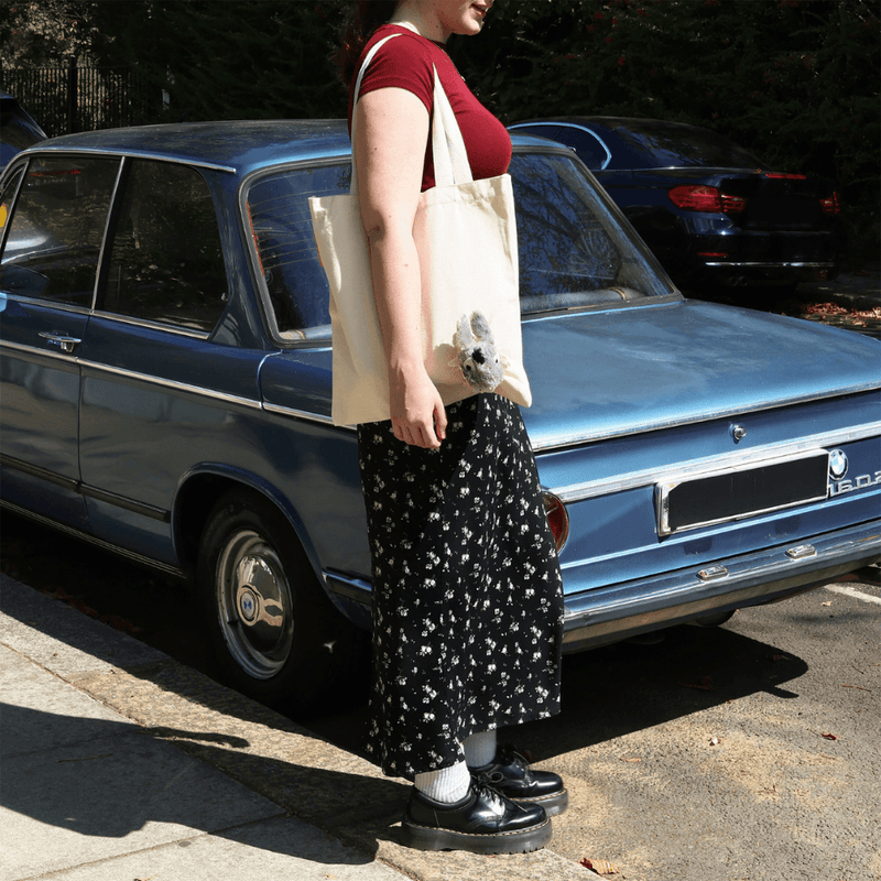 Woman carrying the cream Peeky Donkey tote bag with a plush animal decoration while standing beside a vintage blue car.