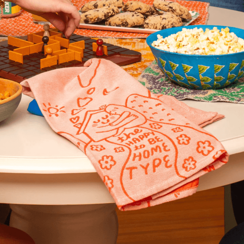 The Happy To Be Home Type tea towel on table, peach woven design, lifestyle photo.