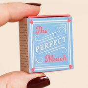 Hand holding The Perfect Match matchbox with blue cover and red heart corners