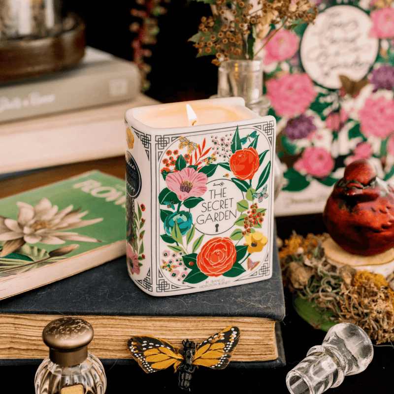 Lit Secret Garden book shaped candle styled on stacked books with floral décor in the background.